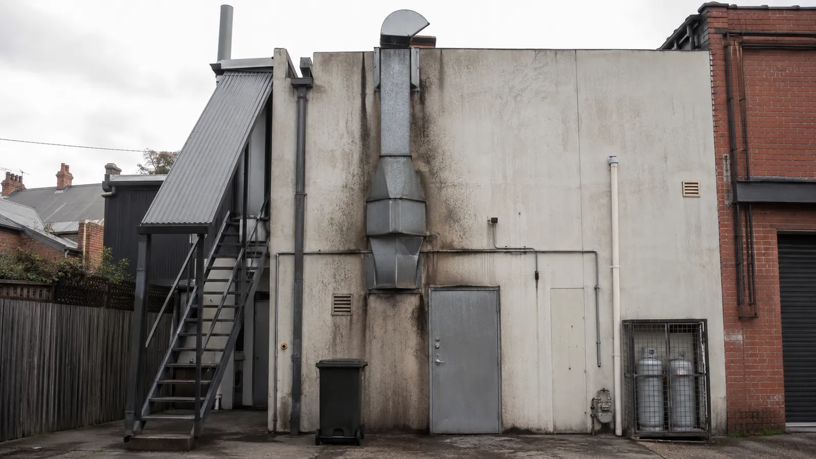 Commercial kitchen exhaust duct beside a rear stair and fire door in a shop-top building service lane