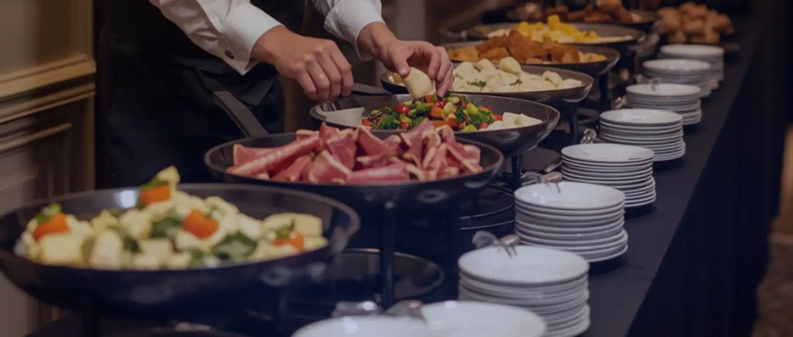 A professional caterer in uniform arranging fresh salads and side dishes on a buffet line next to stacks of clean white plates.