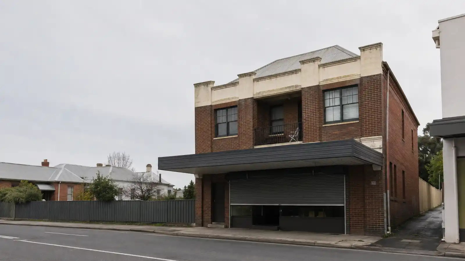 Weathered 1930s brick shop-top building with a half-closed roller shutter and flats above under an overcast sky