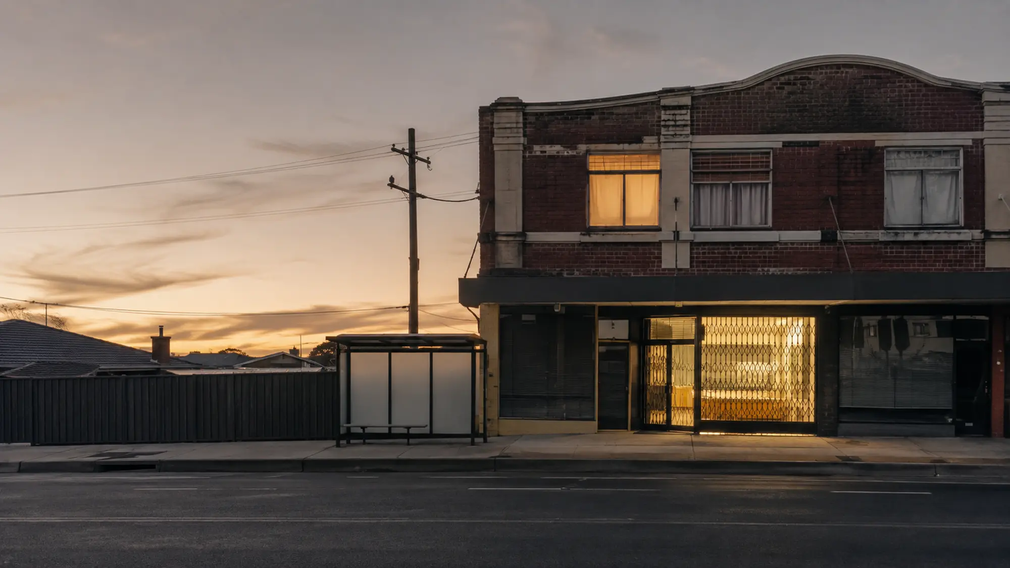 Quiet brick shop-top building with a lit ground-floor tenancy and warm upstairs window at early evening