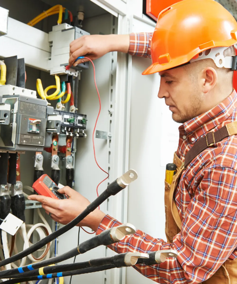 Electrician in hard hat testing industrial switchboard with multimeter for electrical contractor liability insurance