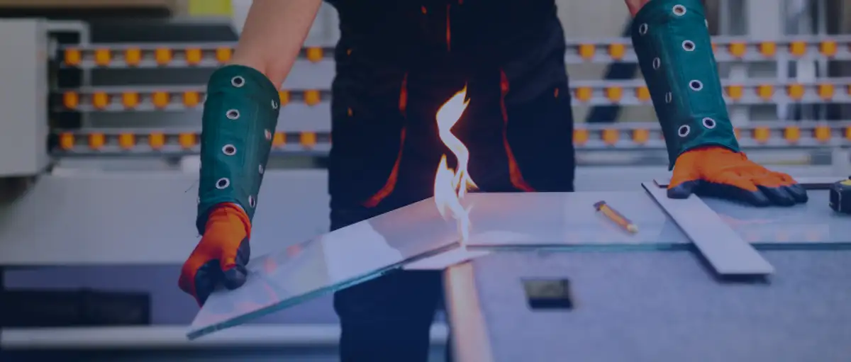 Glazing contractor using flame torch to cut tempered glass sheet while wearing protective arm guards and safety gloves in workshop
