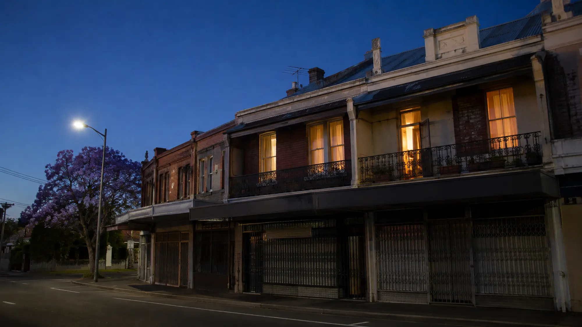 Row of shop-top terraces at dusk with closed shops, warmly lit homes above and a flowering jacaranda