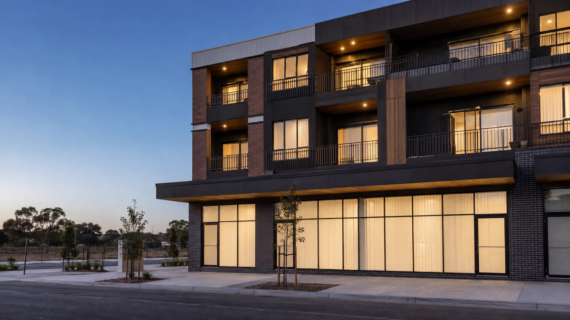 Modern mixed-use building at early evening with lit retail glazing and warm apartment windows